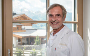 Älterer Arzt in weißem Kittel lächelt vor großem Fenster im malerischen Salzburger Land.