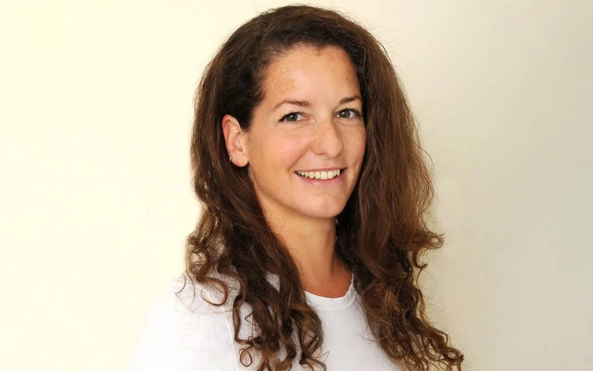 Lächelnde Frau mit lockigem, braunem Haar, vor neutralem Hintergrund.