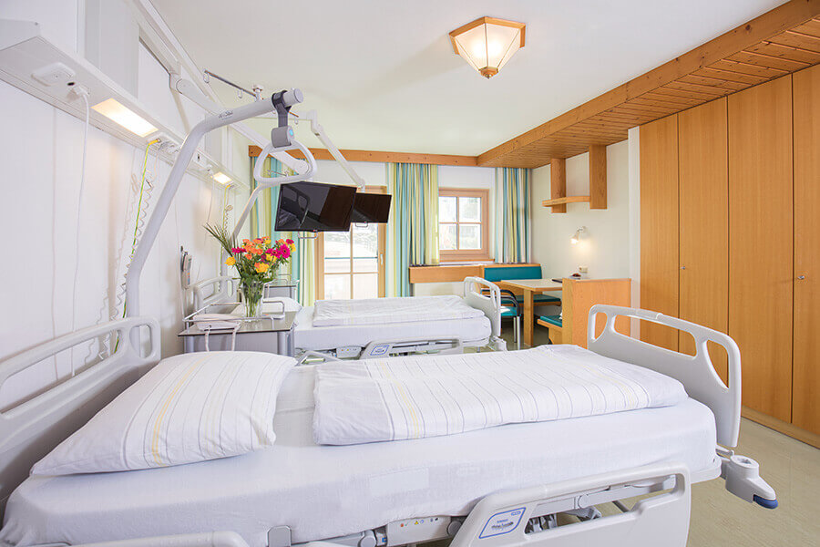 Helle Patientenzimmer mit modernen Betten, TV und einem gemütlichen Sitzbereich.