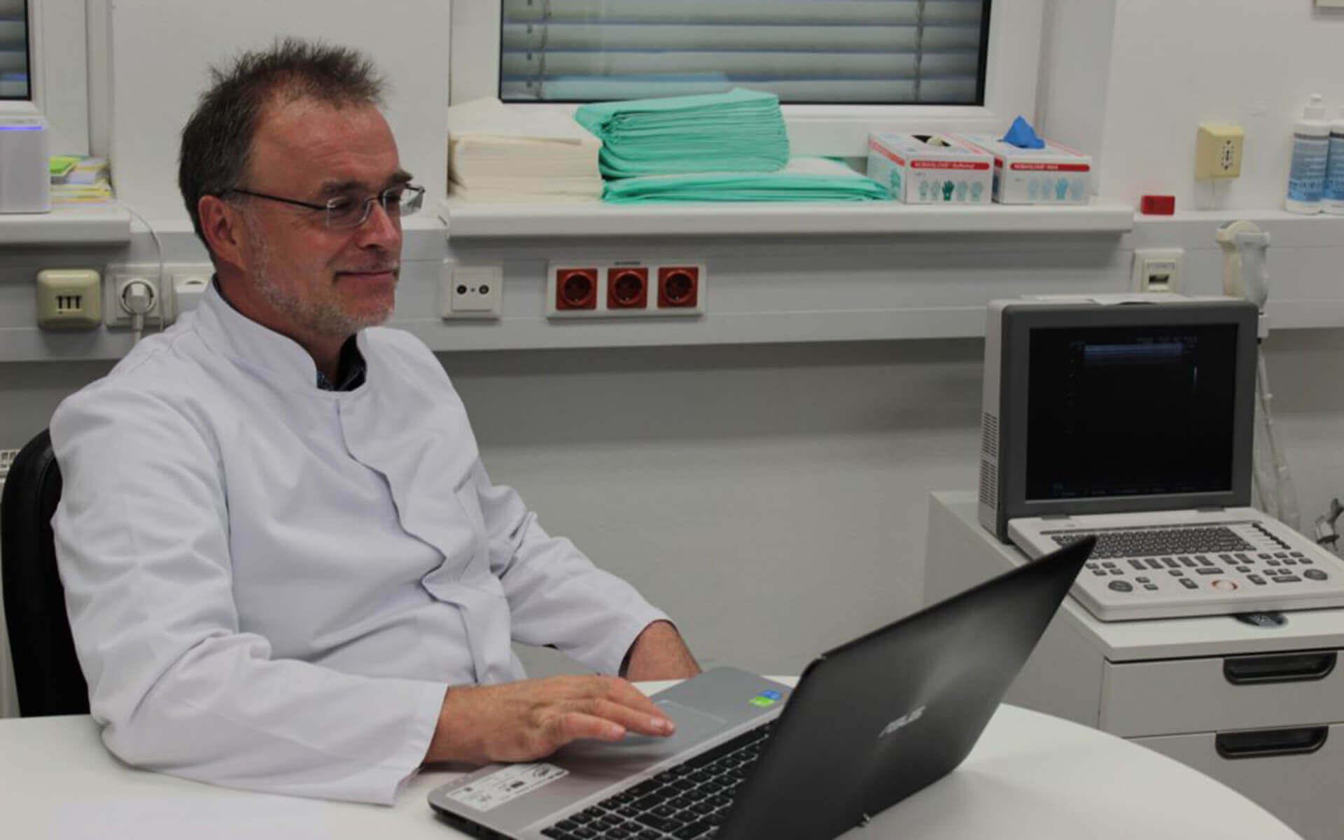 Dr. Peter Metzger in der Klinik, arbeitet am Laptop in moderner Behandlungsumgebung.