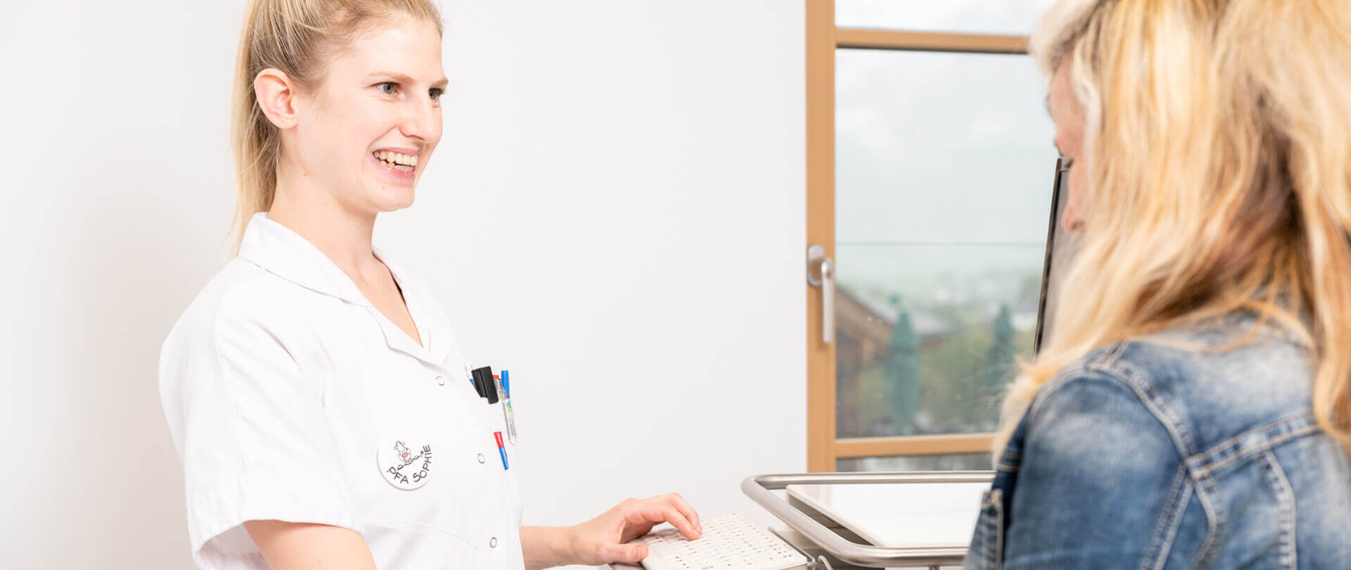 Lächelnde Ärztin im Gespräch mit Patientin in moderner, lichtdurchfluteter Praxis.
