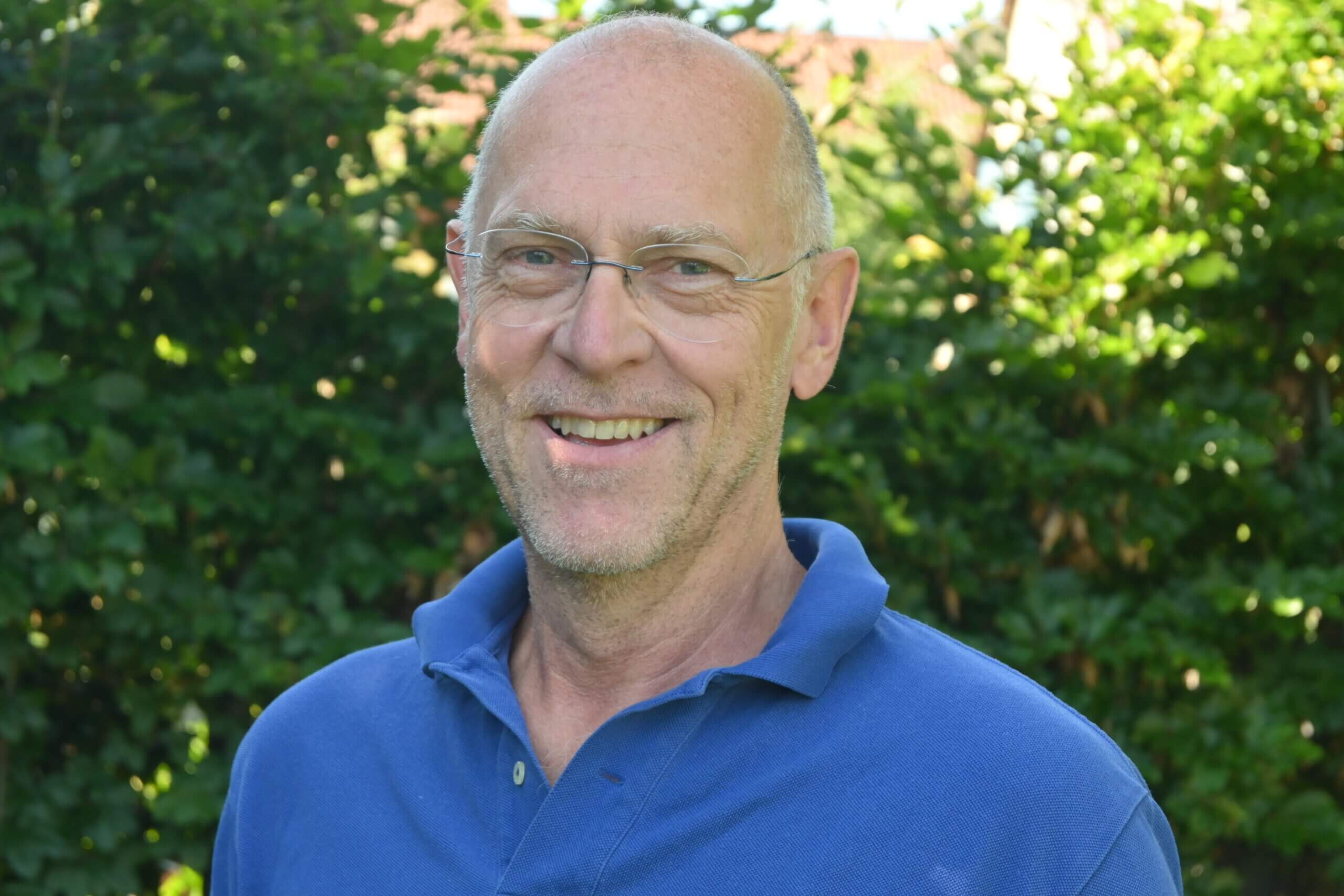 Lächelnder Arzt im blauen Poloshirt vor grünem Hintergrund, vertrauensvolle Ausstrahlung.